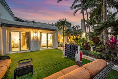 Sunset tropical coastal backyard patio with wicker seating on artificial lawn, sliding glass doors opening to a lit villa, palm trees and a giant four-in-a-row outdoor game.