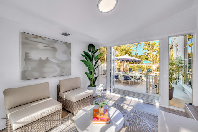 Bright coastal sunroom with wicker seating and neutral cushions, round coffee table with orchid, wide glass doors opening to a sunny patio dining area with umbrella and palm trees