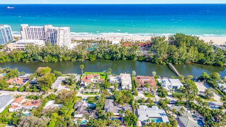 Aerial view of a sunny beachfront neighborhood: palm-lined luxury homes with pools along a calm canal, hotels by a sandy beach and turquoise ocean in the background.