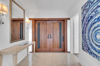Bright modern foyer with solid wood double doors, beige tile floor, minimalist console table and large mirror, accented by a blue circular abstract wall art