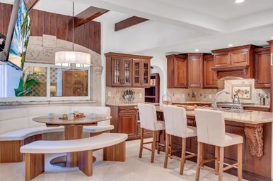 Bright open kitchen with warm wood cabinets, marble island topped by three cream bar stools, curved white banquette breakfast nook with round wooden table and pendant light.