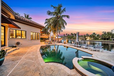 Curved lagoon-style pool and hot tub at a tropical waterfront villa, palm trees and canalfront lounge chairs glowing under a pink-and-blue sunset.