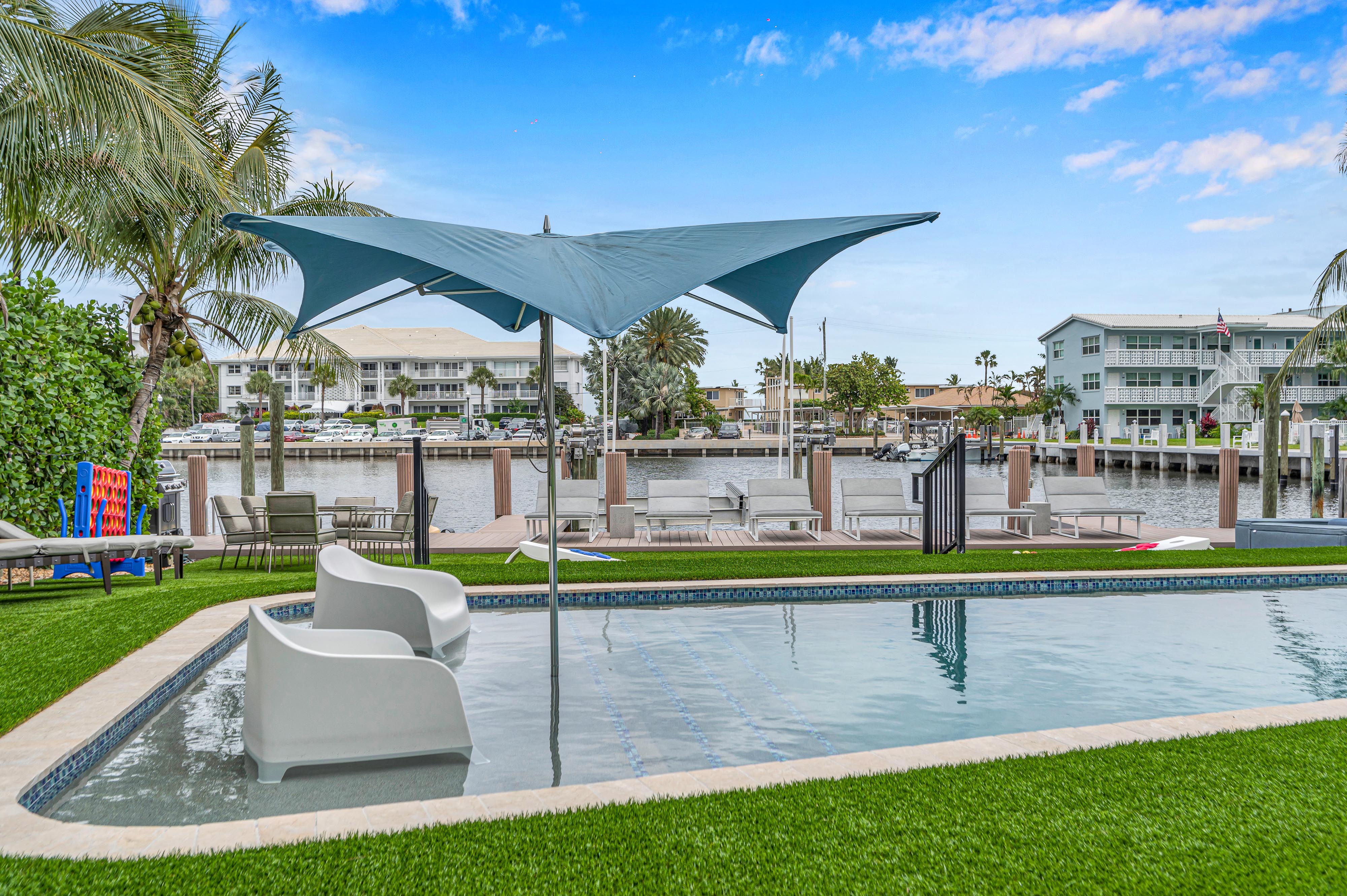 Tropical waterfront pool by a coastal canal with in-pool lounge chairs, blue sail sunshade, palm trees and a wooden dock with boats.