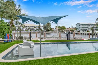 Tropical waterfront pool by a coastal canal with in-pool lounge chairs, blue sail sunshade, palm trees and a wooden dock with boats.
