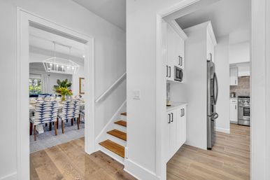 Open-concept home interior with a bright hallway and small wooden staircase, leading to a dining room with blue-and-white patterned chairs and a geometric chandelier, adjacent to a white galley kitchen with stainless steel refrigerator, white cabinets, and wood-look floors.