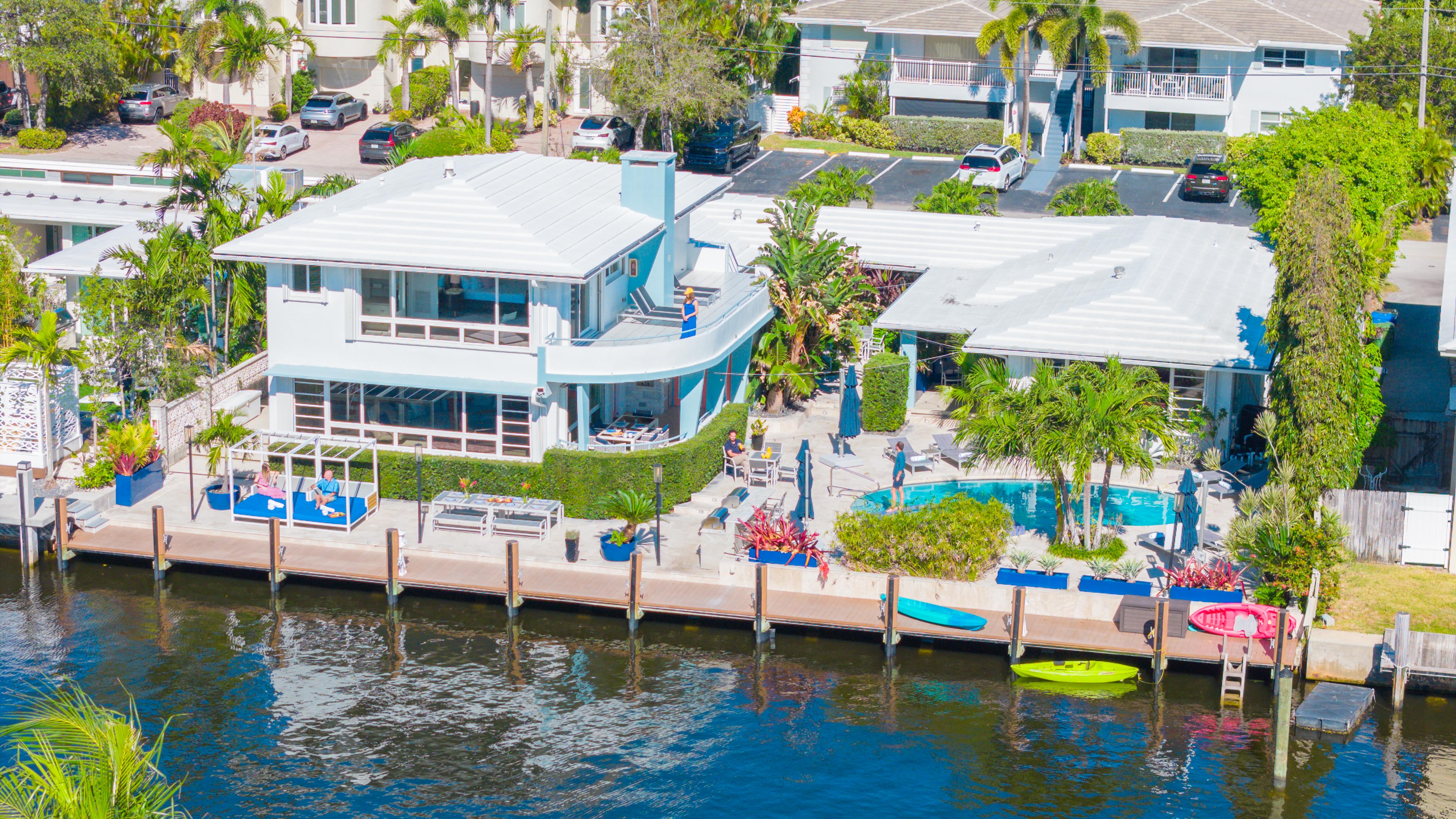 Aerial view of a sunny waterfront canal property with a two-story white house, pool surrounded by palm trees, dockside lounge seating and colorful kayaks tied to the dock.