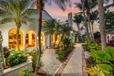 Mediterranean-style home at sunset with arched windows glowing warm, a palm-lined paved walkway, lush tropical landscaping and a decorative tower under a pink twilight sky