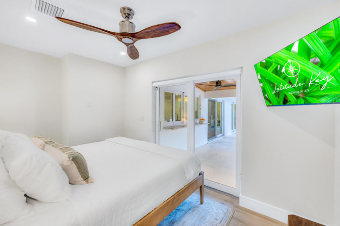 Bright modern coastal bedroom with white linens and wooden bed frame, sculpted wood ceiling fan, wall-mounted TV, and sliding glass doors opening to a covered patio.