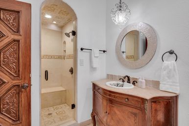 Bright luxury bathroom with a carved wooden door, arched walk-in tile shower with bench, ornate vintage wooden vanity with marble top, round mirror, crystal pendant light and white towels