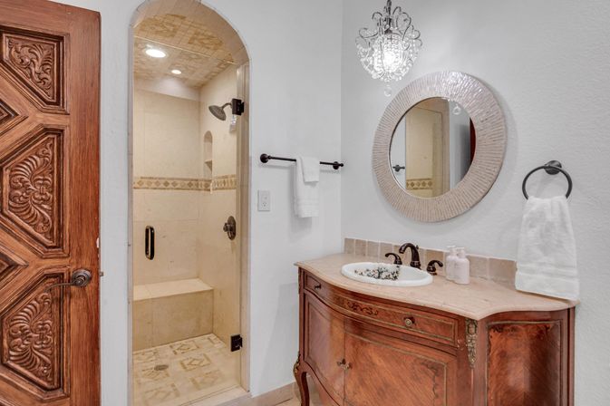 Bright luxury bathroom with a carved wooden door, arched walk-in tile shower with bench, ornate vintage wooden vanity with marble top, round mirror, crystal pendant light and white towels
