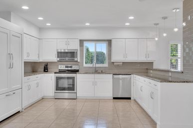Bright modern U-shaped residential kitchen with white shaker cabinets, stainless steel range, microwave and dishwasher, granite countertops, beige subway tile backsplash, pendant lights, tile floor and window overlooking greenery.