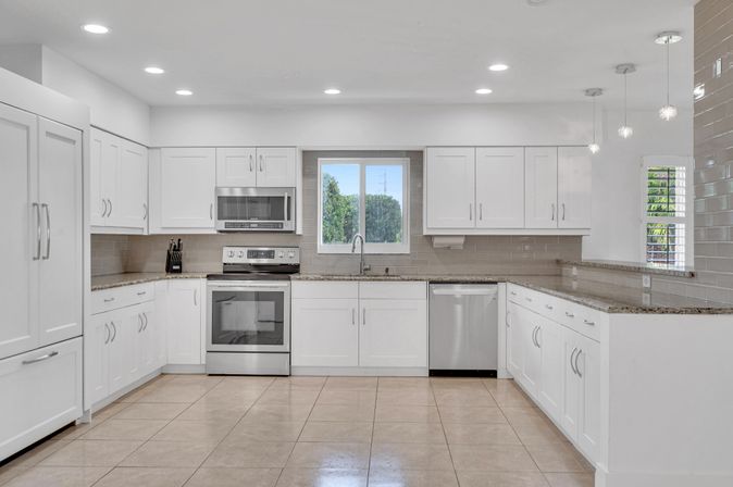 Bright modern U-shaped residential kitchen with white shaker cabinets, stainless steel range, microwave and dishwasher, granite countertops, beige subway tile backsplash, pendant lights, tile floor and window overlooking greenery.