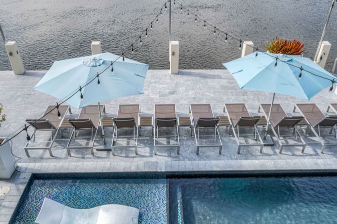 Aerial view of a waterfront pool deck with light-blue umbrellas, a row of sun loungers, hanging string lights and a mosaic-tiled pool beside calm river water.