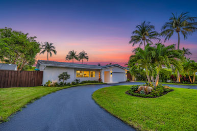Sunset-lit single-story white ranch home with curved driveway, two-car garage, manicured lawn and palm trees creating a tropical suburban vibe.