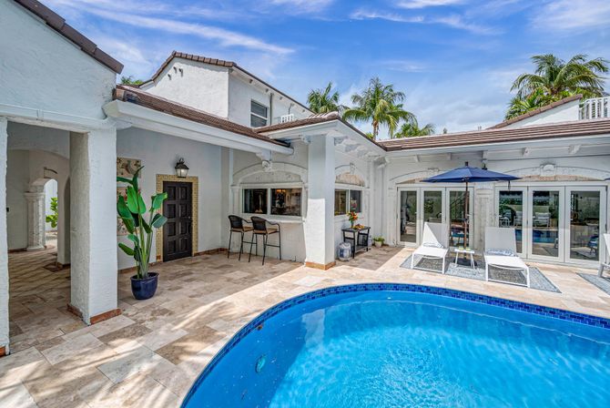 Tropical Mediterranean-style villa courtyard with curved blue swimming pool, tiled poolside patio, outdoor bar with stools, lounge chairs under a navy umbrella, potted plants and palm trees.