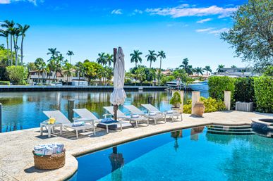 Sun-drenched tropical waterfront pool patio with lounge chairs and umbrella overlooking a calm canal with yachts and palm trees under a bright blue sky