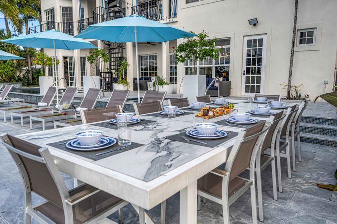 Marble outdoor dining table set with blue-and-white place settings on a poolside patio with lounge chairs, turquoise umbrellas, palm trees and a modern white villa backdrop.
