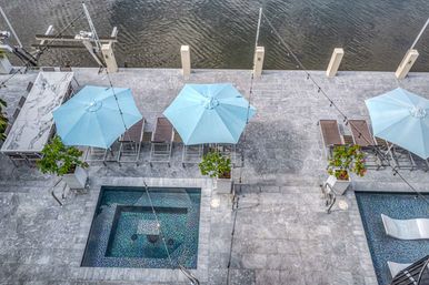 Overhead view of a waterfront rooftop terrace with pale blue umbrellas, lounge chairs, marble dining table, mosaic-tiled plunge pool, potted plants and string lights along the water’s edge.