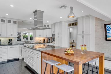 Bright open-concept modern white kitchen with marble-topped island and stainless steel range hood, adjoining wooden butcher-block dining table with bar stools, dark hardwood floors, pendant lighting, decorative 'DINER' sign, and glasses of mimosas.