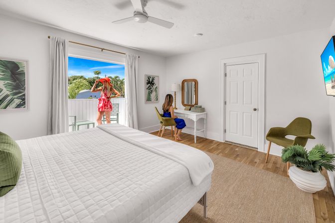 Bright beachside bedroom with white quilted bed, wood floors and jute rug, white vanity and green accent chairs; sliding doors open to a sunny tropical palm view with a person in a red sunhat on the balcony.