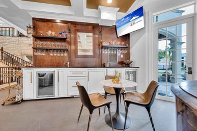 Modern home wet bar with white cabinetry and wood back panel, hanging wine glasses, two beverage fridges, round marble bistro table with three leather chairs, wall-mounted TV and glass doors opening to a poolside patio — bright entertaining space.