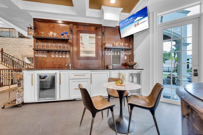 Modern home wet bar with white cabinetry and wood back panel, hanging wine glasses, two beverage fridges, round marble bistro table with three leather chairs, wall-mounted TV and glass doors opening to a poolside patio — bright entertaining space.
