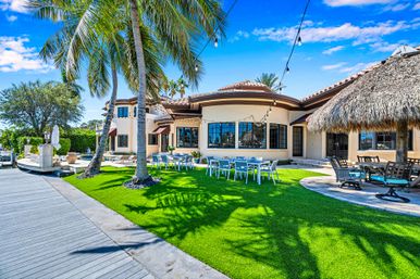 Sunny tropical waterfront villa with palm trees, vibrant green lawn, outdoor dining tables, thatched tiki hut, dock and string lights under a bright blue sky