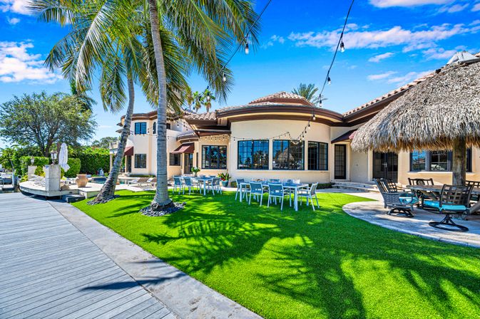Sunny tropical waterfront villa with palm trees, vibrant green lawn, outdoor dining tables, thatched tiki hut, dock and string lights under a bright blue sky