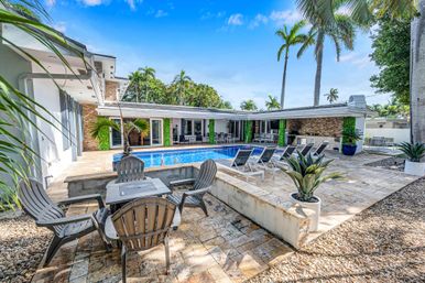 Sunny tropical backyard with a rectangular swimming pool, palm trees, stone patio, lounge chairs, Adirondack seating and an outdoor dining and kitchen area