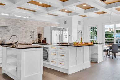 Sunlit luxury white kitchen with large double islands, dark stone countertops, marble subway backsplash, stainless appliances, coffered ceiling, and waterfront dining view.