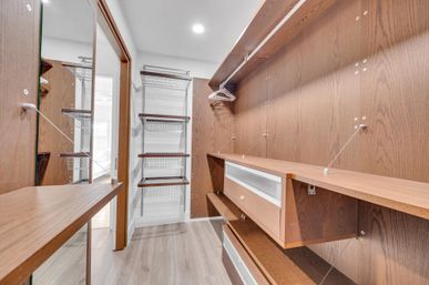 Bright modern walk-in closet with walnut-tone custom shelving and drawers, wire basket racks, hanging rod with hangers, mirrored sliding door, and light wood flooring.