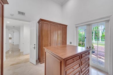 Bright walk-in dressing room with a wooden drawer island and matching armoire, polished stone tile floor, and French doors opening to a tropical patio with palm trees.