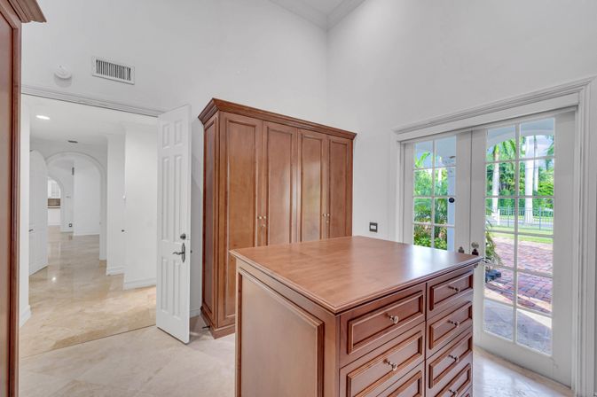 Bright walk-in dressing room with a wooden drawer island and matching armoire, polished stone tile floor, and French doors opening to a tropical patio with palm trees.