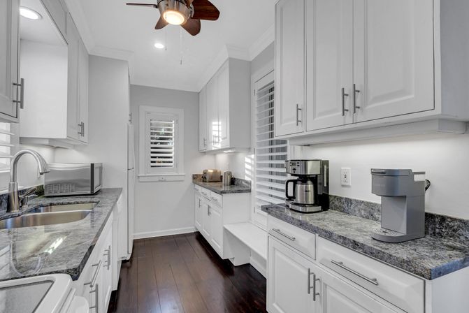 Bright modern galley kitchen with white shaker cabinets, gray granite countertops, dark hardwood floors, stainless sink and microwave, coffee makers and toaster on the counter, plantation-shutter window and ceiling fan