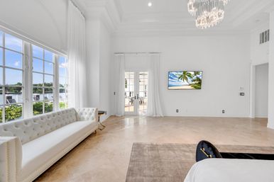 Sunlit waterfront living room with large grid windows, white tufted sofa, marble floors, crystal chandelier, French doors to a patio, and a wall-mounted TV showing a tropical beach scene.