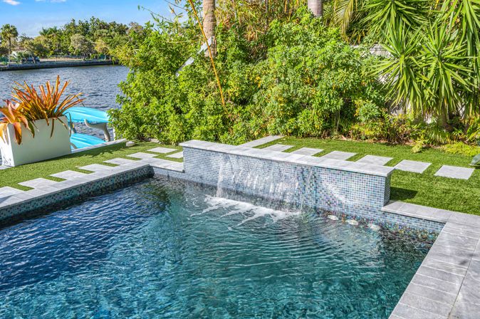 Waterfront tropical backyard pool with mosaic-tiled spillover waterfall, stone stepping pads on green lawn, palms and river view