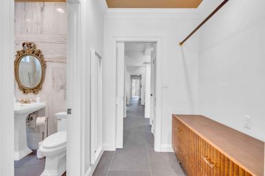 Bright residential hallway opening to a marble bathroom with an ornate gold mirror, pedestal sink and toilet, next to a walk-in closet area with a ribbed wooden storage bench and gray tile floor.