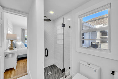 Sunlit modern bathroom with glass-enclosed walk-in shower, black hexagon floor tiles and white subway tiles in a herringbone pattern, white toilet under a window, doorway opening to a bedroom with white bedding and hardwood floors.