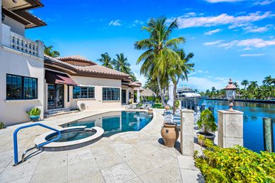 Sunny luxury waterfront villa with curved pool and spa, palm trees along a canal-side patio and a docked yacht under a bright blue sky