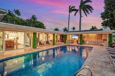 Sunset-lit tropical backyard with a rectangular luxury pool, lounge chairs, covered patio with outdoor kitchen and sliding glass doors, tall palm trees under a pink-purple sky.