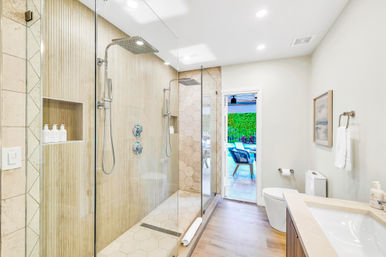 Bright modern bathroom with frameless glass walk-in shower, dual rainfall showerheads, wood-look and hexagon tile, vanity and toilet, doorway to sunny patio with outdoor seating and green hedge