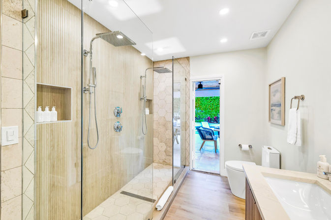 Bright modern bathroom with frameless glass walk-in shower, dual rainfall showerheads, wood-look and hexagon tile, vanity and toilet, doorway to sunny patio with outdoor seating and green hedge