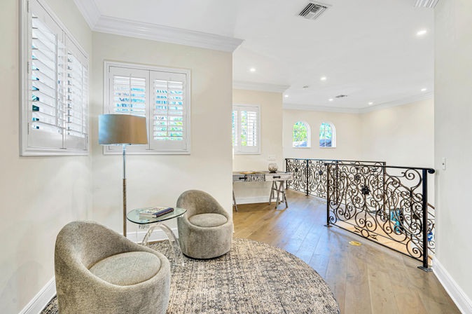 Bright upstairs landing with a cozy sitting area featuring two rounded gray lounge chairs, glass side table and floor lamp on a round textured rug, hardwood floors, white plantation shutters and ornate wrought-iron railing overlooking the staircase.