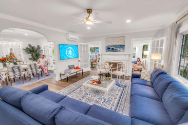 Bright coastal-style open-plan living room with large navy sectional, patterned blue rug, white coffee table with wine and fruit, fireplace topped with seascape art, and glass French doors to a sunlit dining area.