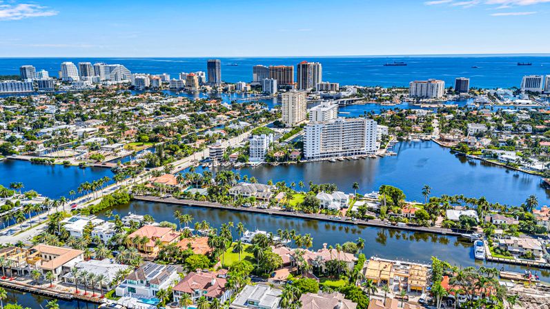 Aerial view of a sunny coastal city with winding canals, palm-lined waterfront homes and yachts, mid-rise skyline and blue ocean horizon.