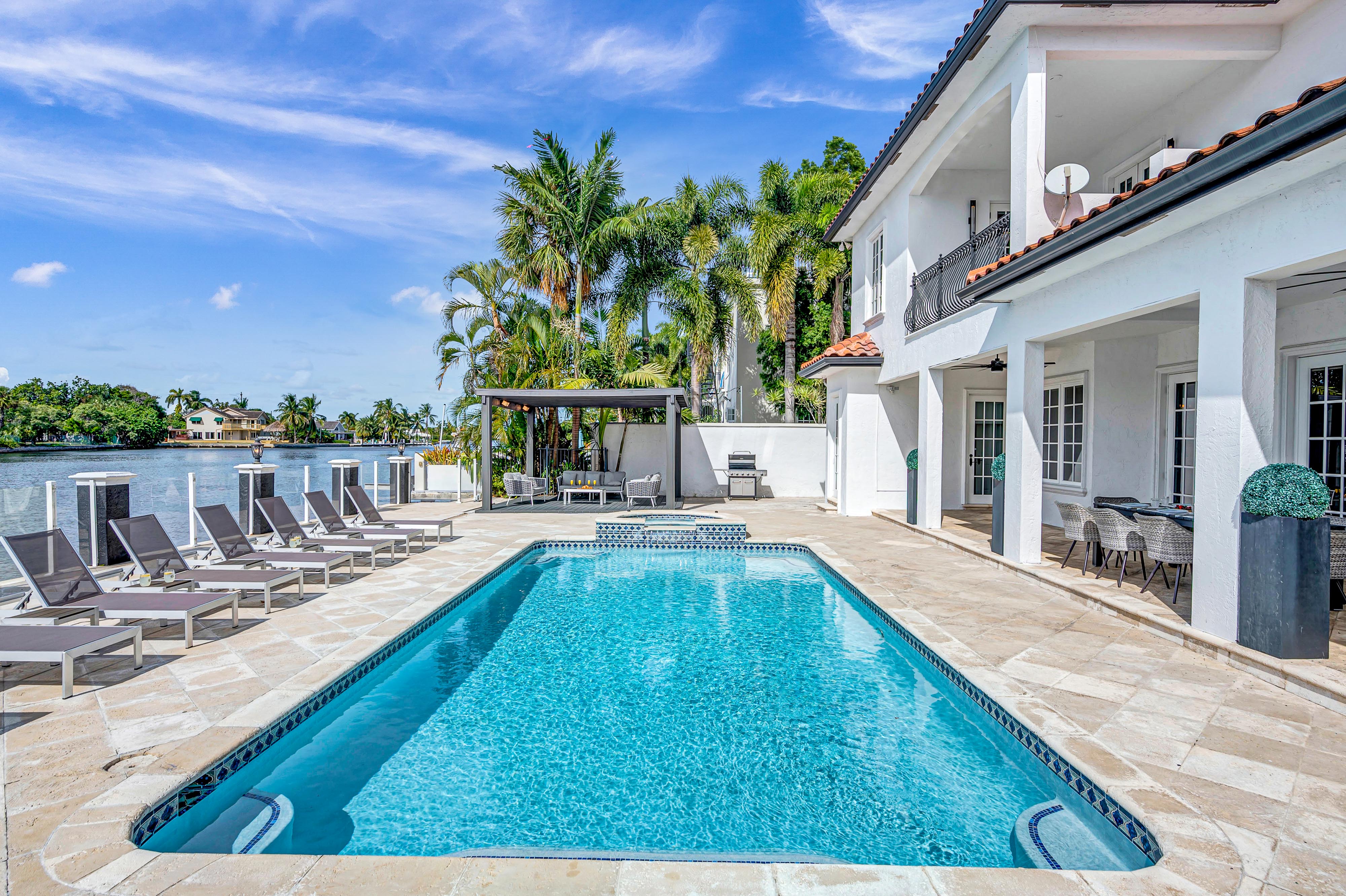 Sunny waterfront pool at a white Mediterranean-style villa with palm trees, lounge chairs, pergola seating and a private dock overlooking a calm blue bay