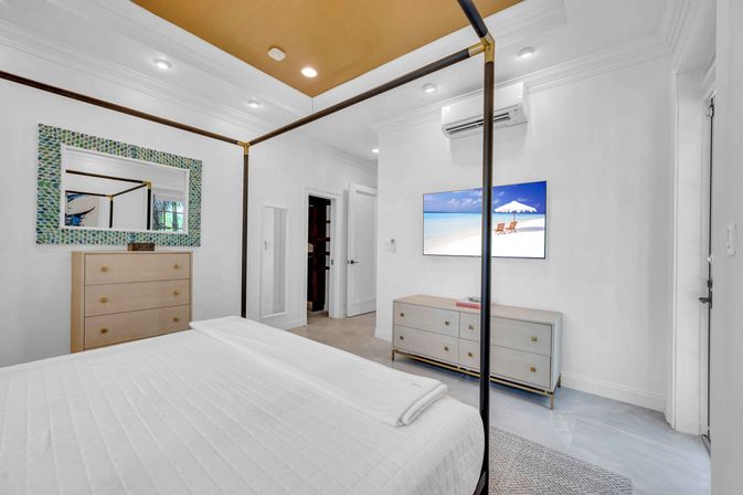 Bright modern master bedroom with a black four-poster bed, white quilted bedding, gold-accent recessed ceiling, wall-mounted TV showing a sunny beach, light wood dressers and a mosaic-framed mirror.