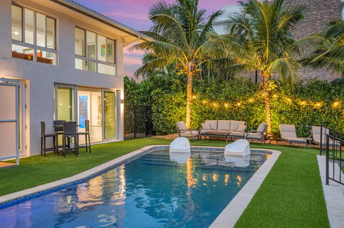 Contemporary backyard pool at dusk with two white floating lounge chairs, palm trees wrapped in string lights, outdoor sofa set on artificial turf and floor-to-ceiling windows of a modern two-story home