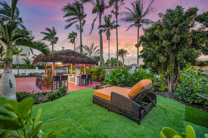 Tropical backyard oasis at sunset with a thatched tiki bar and stools, tall palm trees, lush landscaping and wicker chaise lounges with orange cushions on a manicured lawn