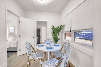 Bright coastal-style dining nook with round white table set with blue dishes, woven bistro chairs, potted palm, hardwood floors, and sunlit windows with roman shades
